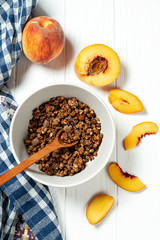Chocolate granola in a white bowl in a composition with a spoon, and peach on white wooden background. Healthy breakfast food. Making breakfast. Gluten free
