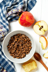Chocolate granola in a white bowl in a composition with honeycombs, a spoon, apple on white wooden background. Healthy breakfast food. Making breakfast. Gluten free