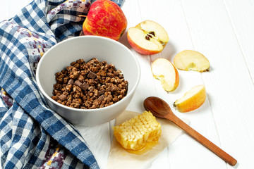 Chocolate granola in a white bowl in a composition with honeycombs, a spoon, apple on white wooden background. Healthy breakfast food. Making breakfast. Gluten free