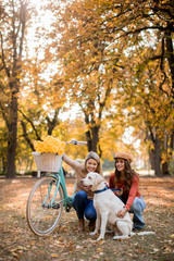 Two female friends walking in the yellow autumn park with dog and bicycle