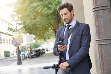 Businessman in the street using smartphone and electric scooter