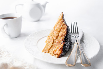 A slice of homemade multilayer biscuit cake. Soaked in butter. Served with chocolate icing and coconut. On a white plate is a fork with a knife. Close-up. White background. 