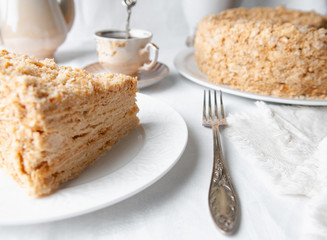 Slice of multilayer Napoleon cake with butter cream. On a white plate. Close-up. Next to a plate is a napkin and a fork. In the background is a cup, teapot and a vase of flowers. White background