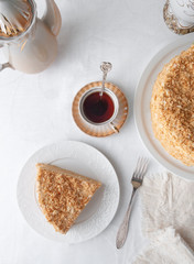 Slice of multilayer Napoleon cake with butter cream. On a white plate. View from above. Near a plate are a napkin, a fork, a cup of tea, a vase of tea with flowers. White background.