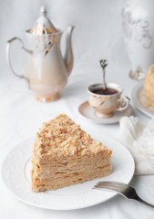 Slice of multilayer Napoleon cake with butter cream. On a white plate. Close-up. Next to a plate is a napkin and a fork. In the background is a cup, teapot and a vase of flowers. White background