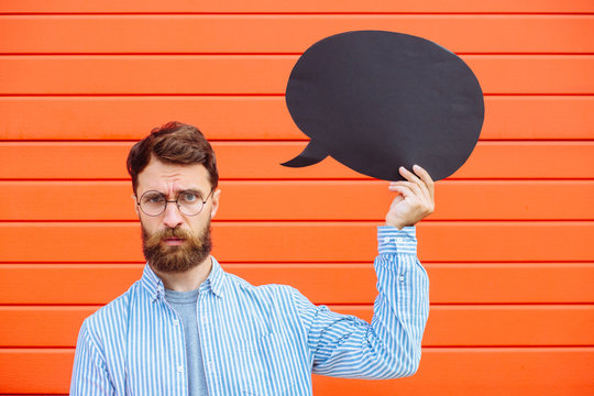 Attractive young beard man in circle glasses with fun fantasy emotion on his face holding a yellow speech bubble looking at camera and smiling, on red background outdoor portrait.