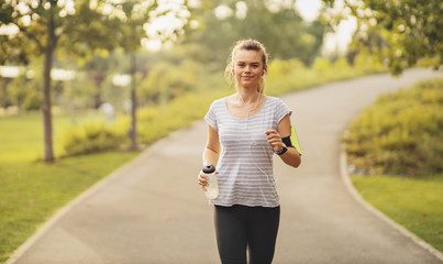 Beautiful fit blonde woman running at the park on a sunny day. Young Female runner listening to music while jogging in the morning.