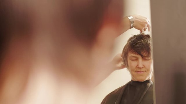 Woman with closed eyes relaxing in front of mirror at hair appointment, hairdresser hands cutting, combing, spraying head in hairdressing beauty saloon, focus on face in glass, then to sprayed hair
