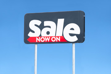Sale now on sign direction arrow to shop against blue plain sky background