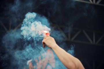 At a rock concert near the stage, a human holds a red signal fire in his hand, which emits thick blue smoke.