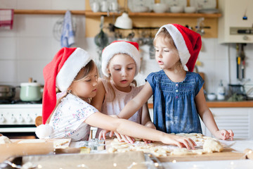 Children are cooking Christmas cookies in cozy home kitchen. Cute kids in santa hats preparation holiday dinner for family. Three sisters bake Xmas biscuits. Lifestyle moment. Children chef concept.