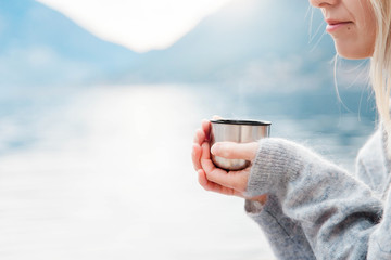 Woman with mug of hot steamy beverage by winter sea, blue mountains. Cozy picnic with thermos of...