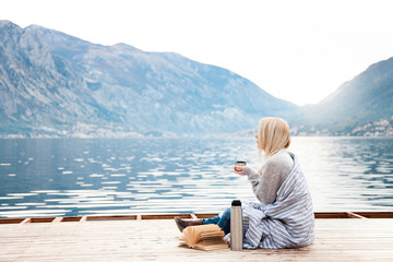 Woman on wooden pier by winter sea, mountains. Cozy picnic with coffee, hot beverages, tea or cocoa...