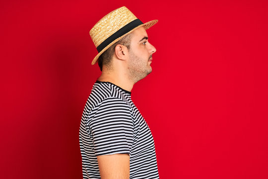 Young man wearing striped navy t-shirt and hat standing over isolated red background looking to side, relax profile pose with natural face with confident smile.