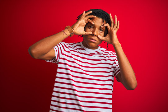 Young afro man with dreadlocks wearing striped t-shirt standing over isolated red background Trying to open eyes with fingers, sleepy and tired for morning fatigue