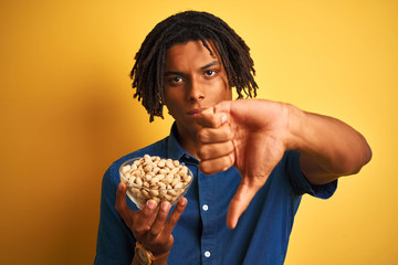 Young afro american man with dreadlocks eating pistachios over isolated yellow background with...