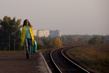 Obraz premium Girl tourist with a suitcase on a railway platform. girl in a yellow raincoat travels. Autumn train ride. Railway in the fall