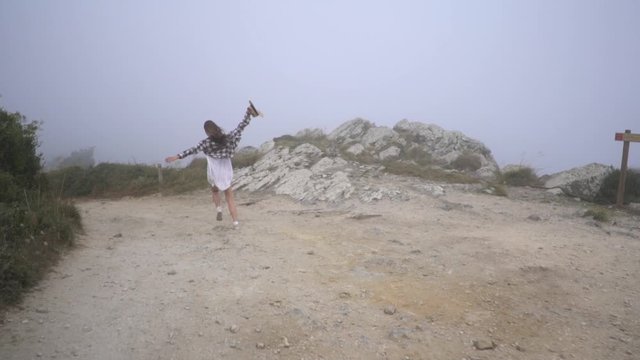 Traveler girl in white dress, plaid t-shirt and straw hat running along country road in fog. Around big stones, plants and trees. Windy, foggy, cloudy weather. Sintra, Portugal.