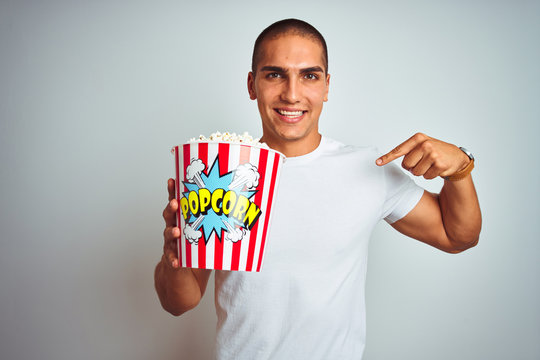 Young handsome man eating popcorn over white isolated background with surprise face pointing finger to himself
