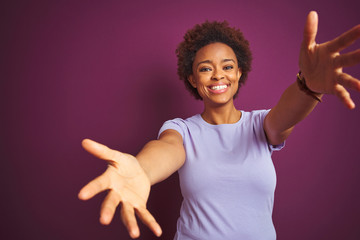 Young beautiful african american woman with afro hair over isolated purple background looking at the camera smiling with open arms for hug. Cheerful expression embracing happiness. - obrazy, fototapety, plakaty