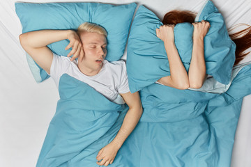 angry girl hiding her head under pillow, trying to sleep, while her boyfriend enjoying sleeping ....