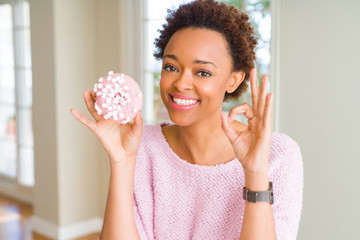 Obraz na płótnie Canvas Young african american woman eating pink sugar marshmallows donut doing ok sign with fingers, excellent symbol