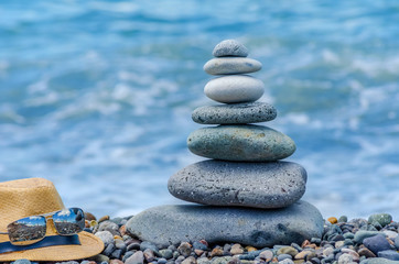 A cairn of stones near the sea, next to a straw hat and glasses. Stone tower on the beach on a sunny day. Balance, peace of mind.