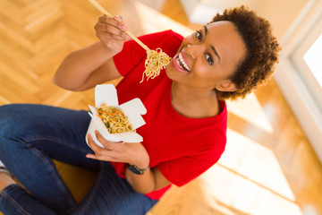 Beautiful young african american woman eating noodles using choopsticks sitting on the floor