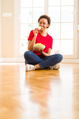 Young beautiful african american woman with afro hair eating chips sitting on the floor at home