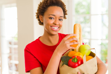 Young beautiful african american woman holding paper bag full of fresh healthy groceries and picking vegetables