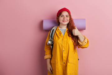 cheerful ginger woman wearing mat a, backback, yellow jacket, cap, shirt hiker shows thumb up, tourits likes her holiday,adventurer expresses positive feeling and emotion. isolated pink background.