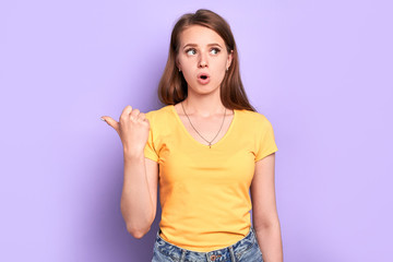 Astonished girl with appealing appearance pointing aside with thumb, showing at some free copy...