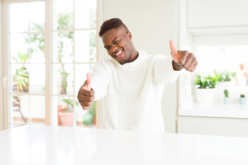 Fototapeta na wymiar Handsome african american man on white table approving doing positive gesture with hand, thumbs up smiling and happy for success. Looking at the camera, winner gesture.