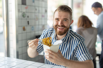 Positive young stylish guy eating chinese noodles in a cafe during a break at work. The concept of rest and healthy snack.