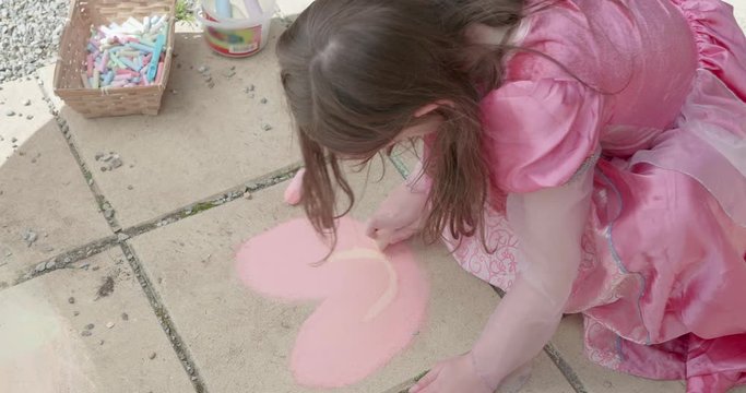 a little girl dressed in a pink princess costume draws a pink heart with a smile in chalk on pavement
