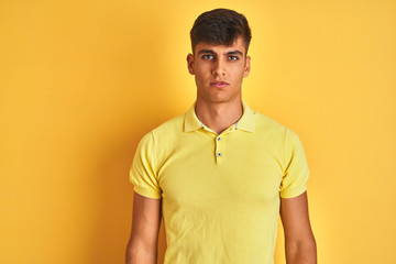 Young indian man wearing casual polo standing over isolated yellow background with serious expression on face. Simple and natural looking at the camera.