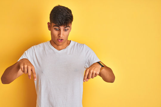 Young indian man wearing white t-shirt standing over isolated yellow background Pointing down with fingers showing advertisement, surprised face and open mouth
