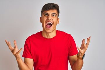 Fototapeta premium Young indian man wearing red t-shirt standing over isolated white background crazy and mad shouting and yelling with aggressive expression and arms raised. Frustration concept.