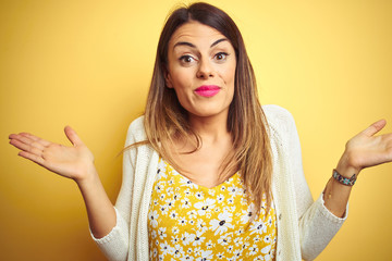 Young beautiful woman wearing jacket standing over yellow isolated background clueless and confused expression with arms and hands raised. Doubt concept.