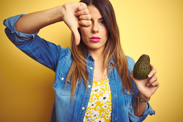 Young beautiful woman eating healthy avocado over yellow background with angry face, negative sign...