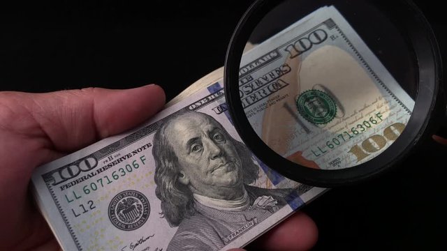A man checks dollars through a magnifying glass. In the man&rsquo;s hands is a magnifying glass and one hundred American dollars. Checking banknotes with a face value of one hundred dollars for a fake.