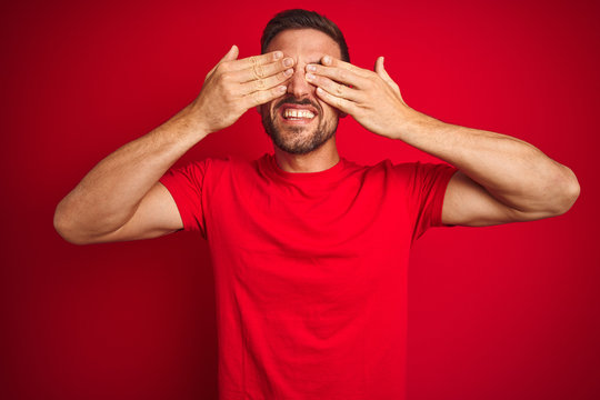 Young handsome man wearing casual t-shirt over red isolated background covering eyes with hands smiling cheerful and funny. Blind concept.