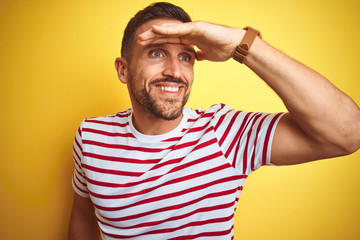 Fototapeta premium Young handsome man wearing casual red striped t-shirt over yellow isolated background very happy and smiling looking far away with hand over head. Searching concept.