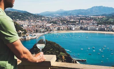 Fototapeta premium Drink glass white wine in male hands holidays looks top view city coast yacht from observation deck, tourist man toast alcohol panoramic cityscape downtown, tourism san sebastian vacation enjoy travel