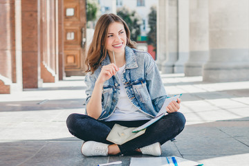 Girl student sits opposite the university and smiling. Cute girl student holds pensil, folders, notebooks and laughs. Girl teaches lessons