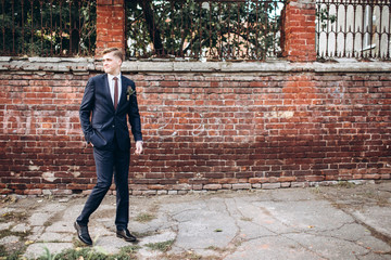 Fototapeta na wymiar brutal groom in a suit and tie. The blond guy is walking around the old town. Young male businessman on brick wall background. Portert of a smiling guy.