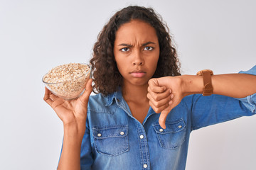 Young brazilian woman holding oat bowl standing over isolated white background with angry face,...