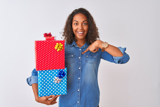 Young brazilian woman holding birthday gifts standing over isolated white background with surprise face pointing finger to himself