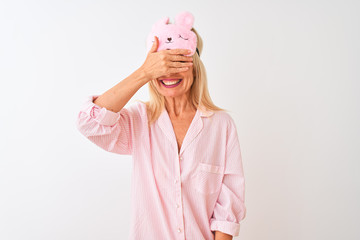 Middle age woman wearing sleep mask and pajama over isolated white background smiling and laughing with hand on face covering eyes for surprise. Blind concept.