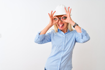 Middle age architect woman wearing glasses and helmet over isolated white background doing ok gesture like binoculars sticking tongue out, eyes looking through fingers. Crazy expression.